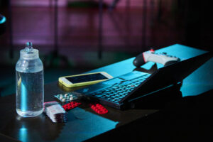舞台セットの机の上に乗っている、ボトル、薬、スマートフォン、PC、ゲームコントローラー