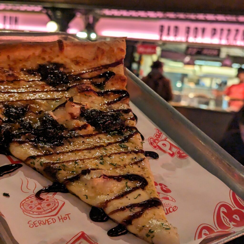 バルサミコ酢がかかった薄めの一切れのピザ。後ろにはピザ屋の看板が見える。